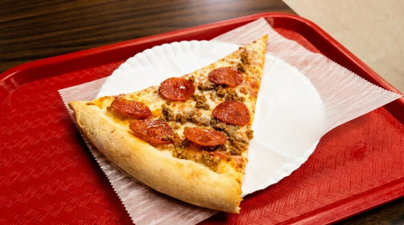 A single slice of pepperoni and sausage pizza on a white paper plate, resting on a red tray.